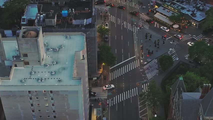 People walk along the busy streets of New York City as cars move through intersections. Shops and street lights glow in the evening light, creating lively scenes.