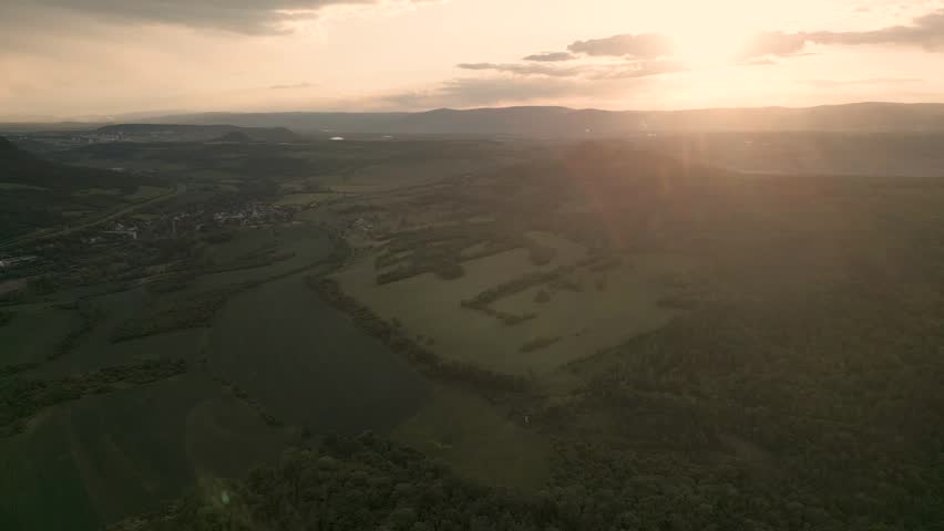 An aerial panorama captures the rugged, vertical basalt cliffs of  hill rising dramatically above a misty autumn landscape. Dense green forests surround the base of the peak, while soft sunlight filte