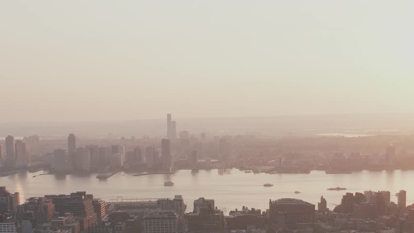 The skyline of New York City is visible at sunset. Buildings reflect the warm colors of the sun. The river flows gently as boats move along the water.