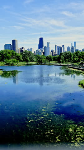 A park features a lake surrounded by green trees with a view of the Chicago skyline in bright daylight. The buildings rise above the landscape, showcasing the city.