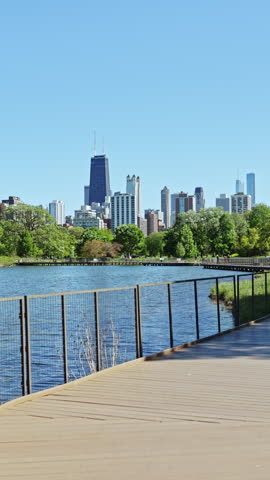 View shows a city skyline behind a lake with green trees. People stroll along a wooden walkway beside the water under a clear blue sky.