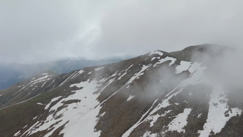 Snow-covered mountain peaks with rocky terrain are partially obscured by fog, creating a dramatic landscape. The scene captures the natural beauty of a mountainous region under cloudy skies