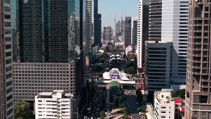A view of Bangkok with tall buildings and busy streets. People move around while vehicles travel along the roads. Construction is visible in the background.
