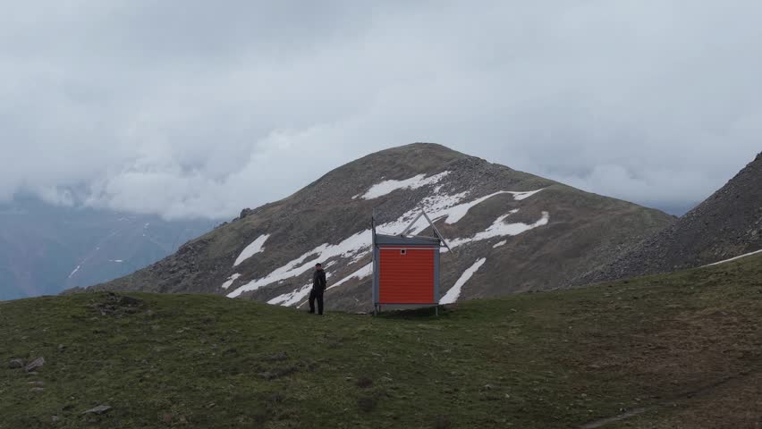 A hiker in dark clothing walks towards an orange shelter on a grassy mountain slope, with snow-capped peaks and cloudy skies creating a dramatic backdrop for the scene