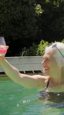 Vertical video: Senior pushing glass across pool, blue-swimsuit woman reaching for drink. Outdoor, backyard, water, sunglasses, lounge, relaxation, vertical video