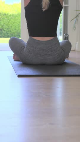 Vertical video: Stretching woman in gear, sun shining, moving into meditation on mat by glass doors. Yoga, home, calm, serenity, minimalist, daylight, vertical video