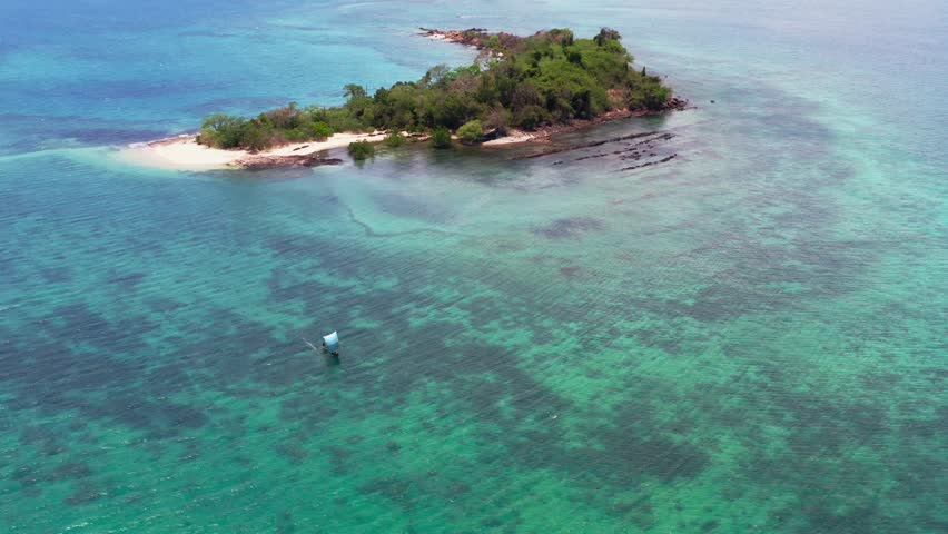 Scenic aerial drone view of a traditional sailboat cruising near a lush tropical island surrounded by clear turquoise coral reefs in Nosy Komba, Madagascar.