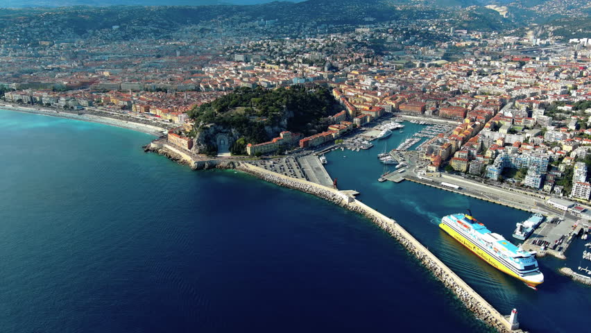 High angle aerial drone footage shows a large yellow ferry entering Port Lympia in Nice, France, next to Castle Hill and the Mediterranean coastline on a clear sunny day.