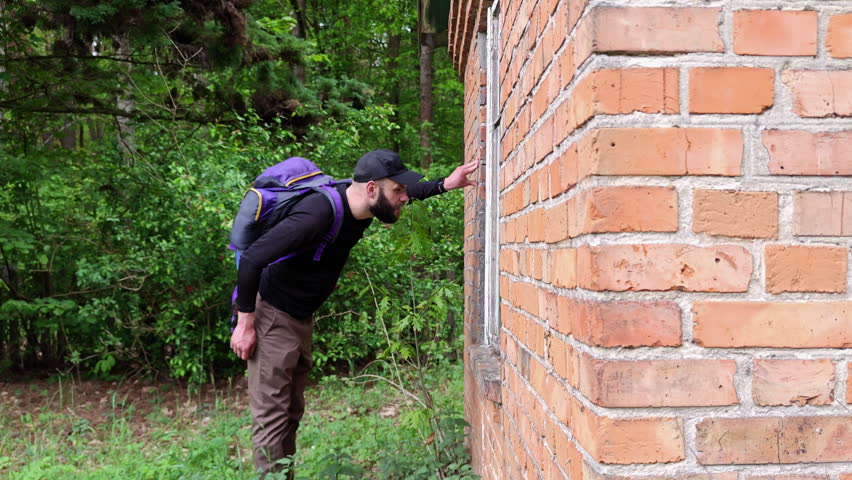 Hiker with a backpack looking inside the window of an old brick house from the outside