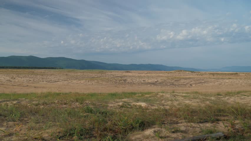 Handheld static shot of a dry, yellow grass field in Russia. The scenic landscape features a sandy midground leading up to green mountains under a bright, cloudy blue sky.