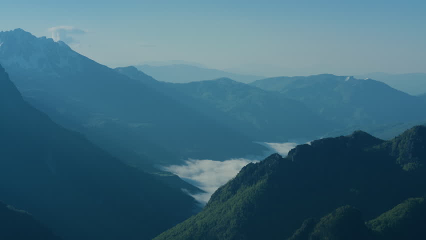 Mountain valley with low clouds and blue mist, static top view landscape. Snowy alpine peaks under a clear sky overlooking a beautiful green meadow.