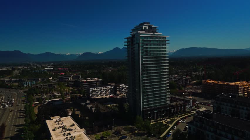 Two modern glass residential towers rise above sprawling suburban district revealing expansive highway network commercial buildings distant snowcapped mountain peaks beneath clear bright blue sky