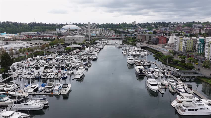 Hundreds of boats fill Thea Foss Waterway marina with Tacoma Dome and cable-stay bridge rising beyond colorful waterfront apartment buildings in Tacoma, Washington, USA. Aerial drone flyover shot,