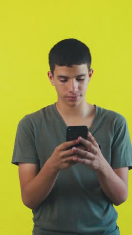 Handsome hispanic teenager happily texting on his mobile phone. Young man smiling while using a social media app on a yellow background