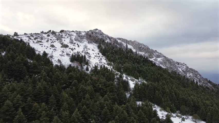 Scenic horizontal drone shot flying over a pine forest towards the snow dusted rocky ridge and communication towers of Vounasa peak in Greece. High quality 4K winter footage.