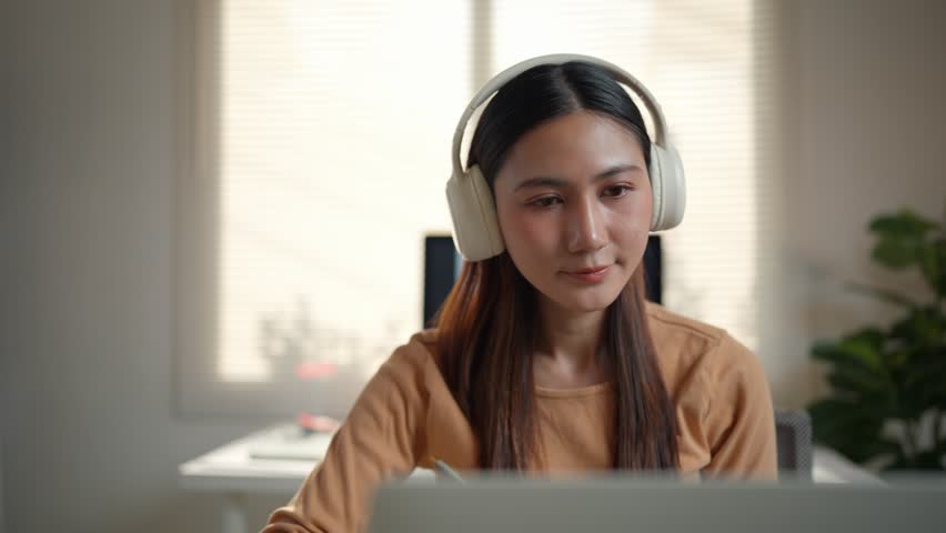 Focused Asian woman wearing headphones, learning and taking notes while attending a university webinar. Online education and e learning concept