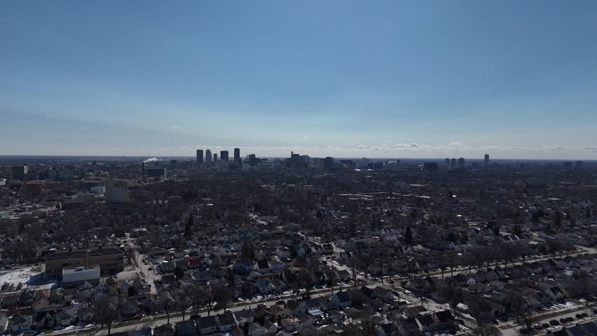 Winnipeg, Manitoba residential area. Vertical drone rises straight up revealing street grid, suburbs, and downtown skyline under sunny spring skies with distant clouds.