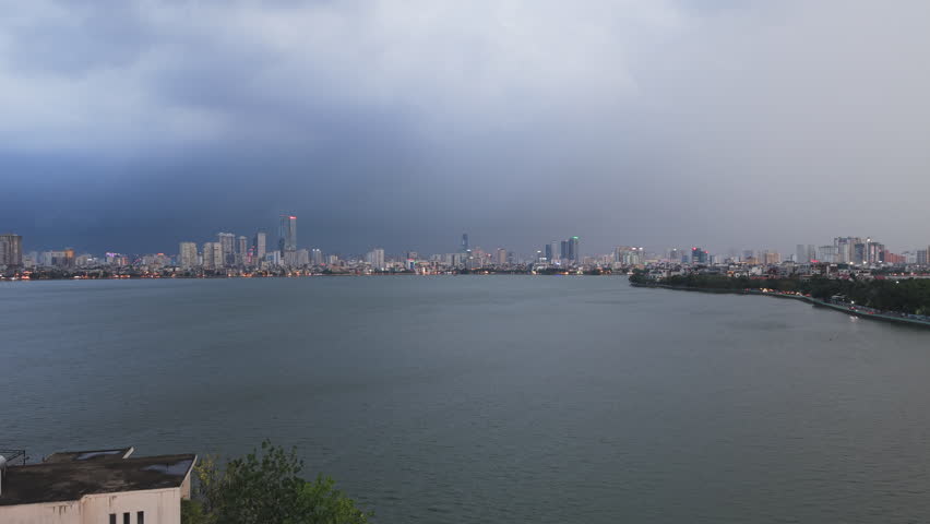 Slow drone push over west lake toward Hanoi skyline under heavy storm clouds at dusk, climate of Vietnam city.