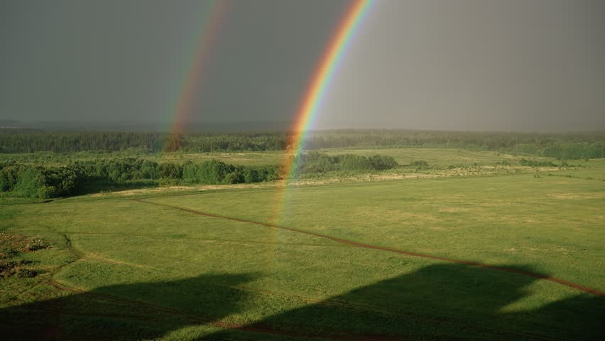 Stunning aerial footage capturing a bright double rainbow forming above a vast green field and forest after rain, with its colorful arc gradually fading into the overcast sky
