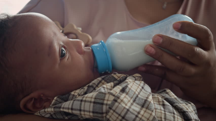African American infant drinking milk from bottle held by mother in close-up shot. Ideal for baby formula advertisements, parenting blogs and work-life balance