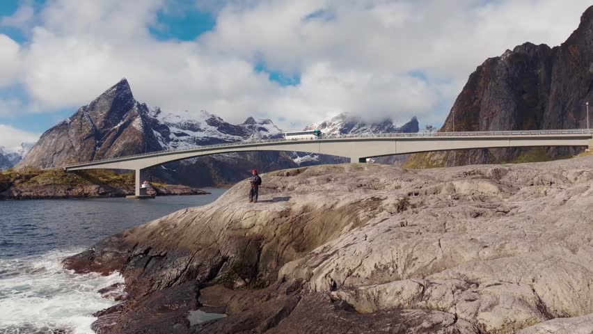 Person on rocky shore facing arched concrete bridge and snow capped mountains