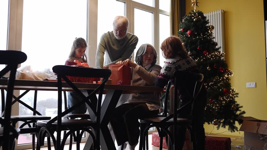 Grandmother sits at a table and wraps a gift while talking to her grandson and grandfather holding scissors