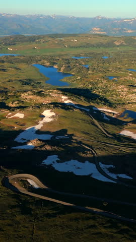 Vertical Screen: Beartooth Highway in Montana USA with winding mountain road cutting through alpine terrain with snow patches and blue lakes dotting the green valleys below.