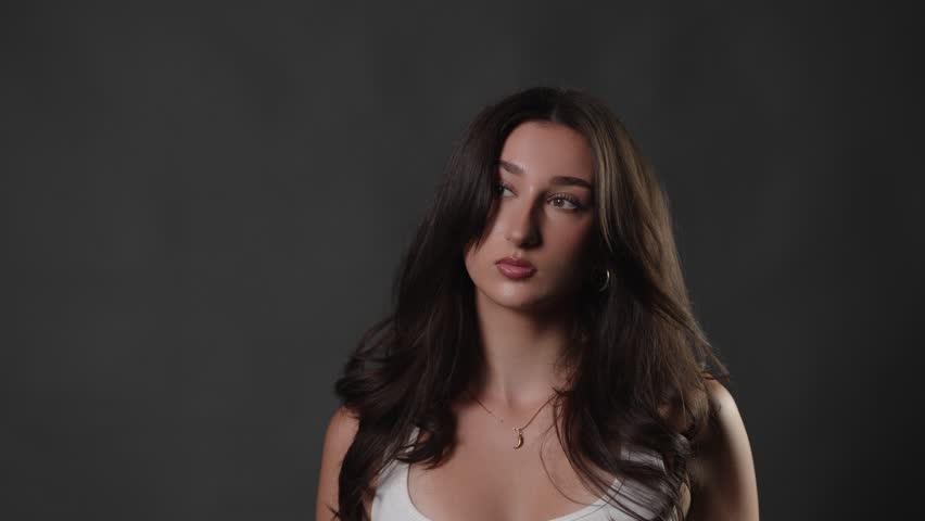 Beauty and fashion portrait of a young woman with long dark hair posing in a studio, embodying natural beauty and confidence. The model a serene expression on a neutral gray background, highlighting h