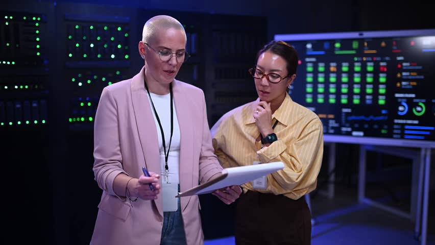 Two diverse female it specialists discussing network maintenance and data analysis in a high tech control room with server racks