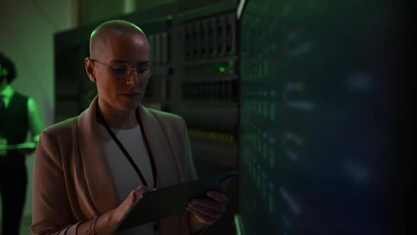 Young woman with glasses and a shaved head working in a data center. An it specialist analyzing network data on a tablet
