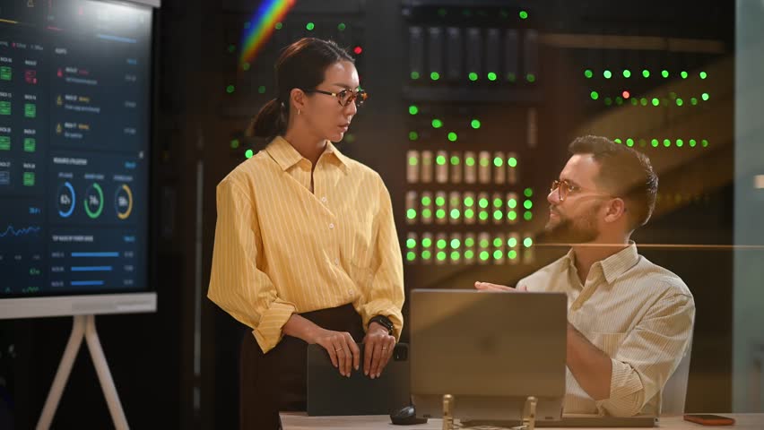 Asian woman and caucasian man discussing server performance in data center. Colleagues working with laptop in modern server room