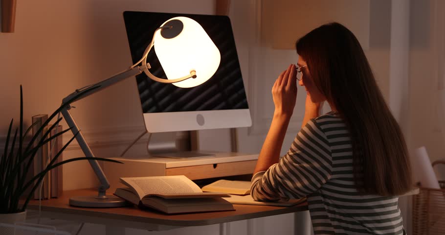 Tired woman at wooden desk with lamp and computer monitor indoors