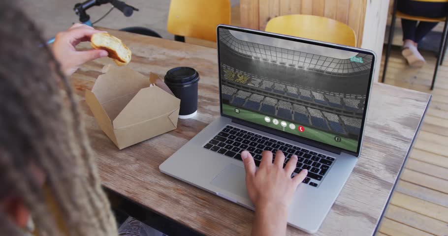 Man working on laptop showing map overlay at cafe while reaching takeout box and eating pastry. Remote, globe, typing, device, coffee, bicycle, wooden