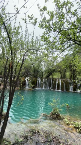 Crystal clear turquoise water cascades over multiple waterfalls, framed by lush green trees and foliage in the serene Jiuzhaigou Valley. This UNESCO World Heritage site in Sichuan, China, is