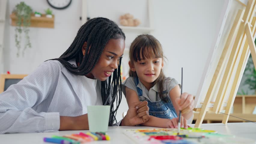 African woman tutor guide young child through art and painting in nursery. Caring female teacher offers support and encouragement to adorable little girl creating artwork in school education center.