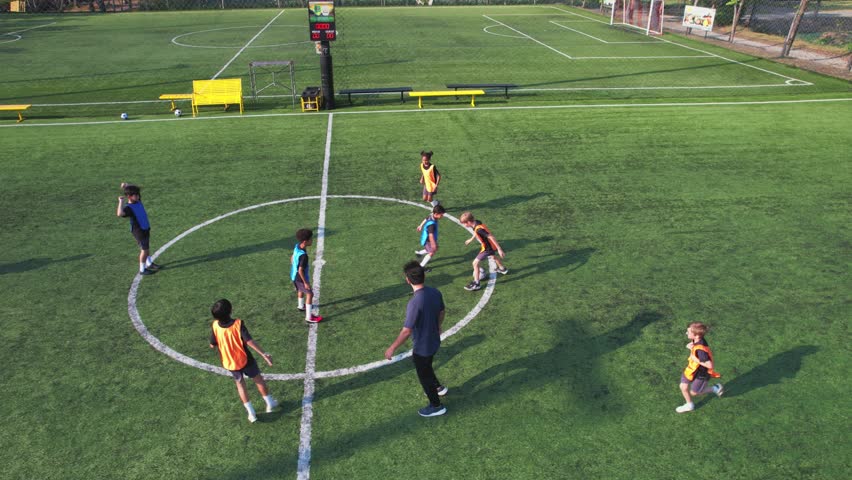 Aerial view of children practicing football on outdoor field with coach. Group of young active kid players practicing soccer skill and teamwork with trainer support during outdoor training activity.