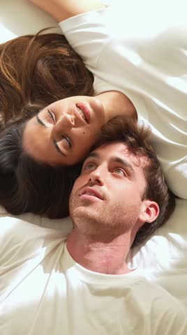 Young heterosexual couple in white t-shirts smiling and relaxing while lying in bed, enjoying a tender moment together and looking at the camera from a top-down perspective