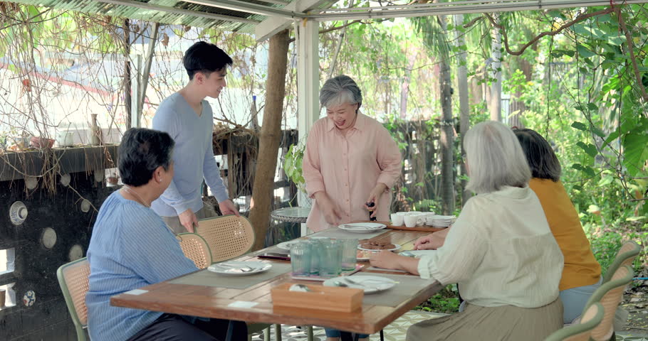 Senior Asian women friend reunion at garden restaurant, young man arriving at brunch table while retired friends welcome family member, sharing togetherness, social connection, warm multigenerational moment.