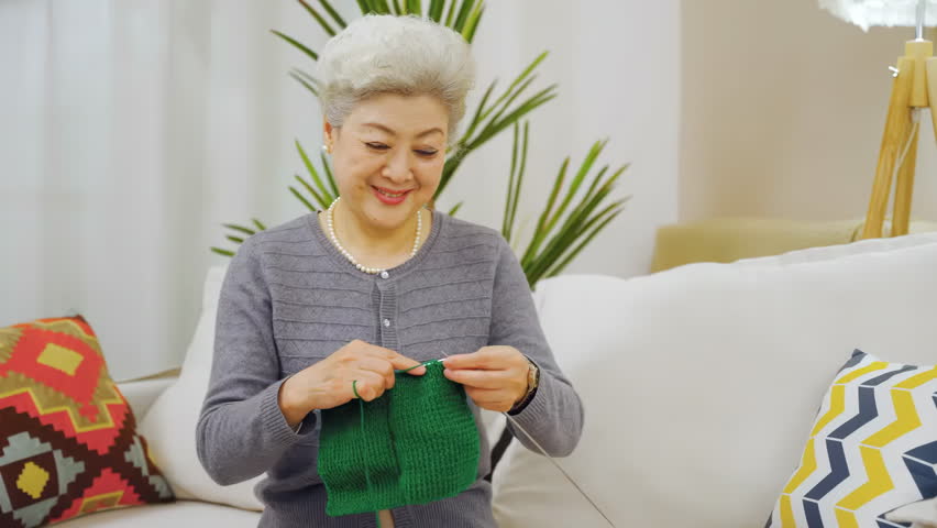 Smiling skilled elderly grandmother woman knitting green sweater by yarn ball and needles with wrinkly hands crafting hobby in free time sitting comfortably on sofa couch. People, age and leisure.