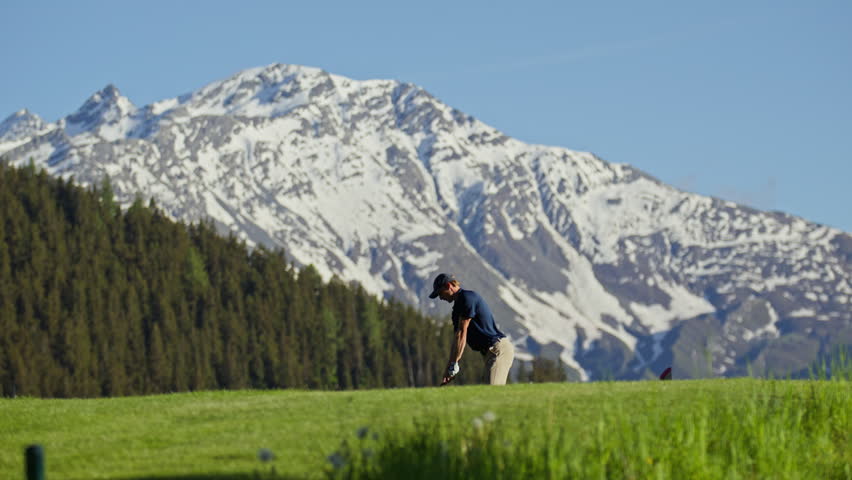Professional golfer taking a full swing on the fairway at Verbier Golf Club in Switzerland. The snow-covered peak of Le Rogneux rises above the alpine course, creating a spectacular mountain golf sett