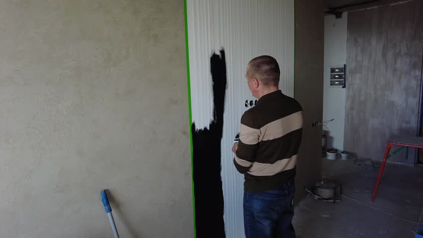 A person is painting a wall black in a room of a building. The wall is unfinished and has a smooth texture. The painter is using a roller brush and seems focused on the task. There is a green tape along the edge of the wall, helping to create a straight line. Various tools and materials are visible in the area, indicating that the space is still under renovation. Light from the windows helps illuminate the room as work continues.