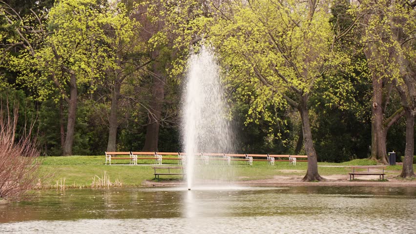 Water fountain sprays upward from a park pond as ripples move across the water, with trees and benches visible in a static wide shot.