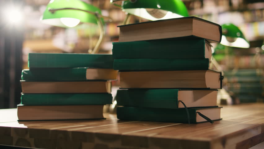 Stack of books on desk in quiet university library with warm lighting and green lamp, study materials and educational environment with wooden bookshelves.