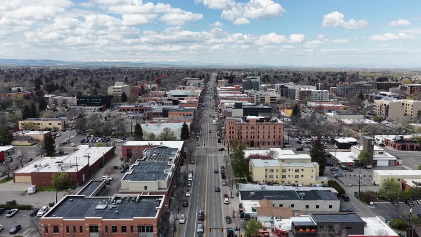 Downtown Bozeman Main Street stretches toward distant mountains across flat valley with brick storefronts, solar rooftops and spring trees under blue skies, Montana, USA. Aerial drone rising wide shot