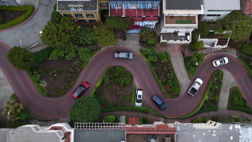 Lombard Street winding brick curves lined with lush manicured garden beds as cars slowly navigate famous hairpin turns in San Francisco, California, USA. Aerial drone top down shot. Famous scene.