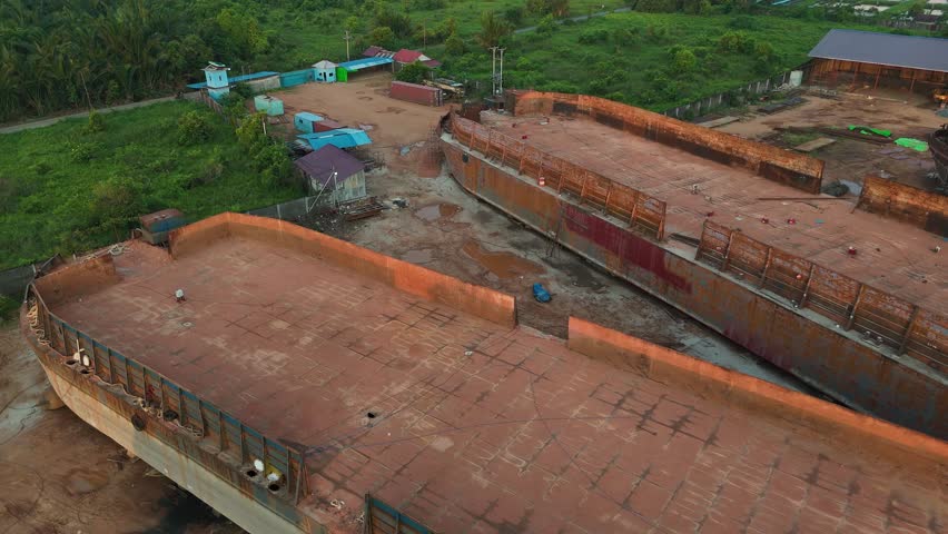 An aerial view showing a dry dock with two large, rusty steel barges side by side at the shipyard, with a crane and an old ship in the background. 