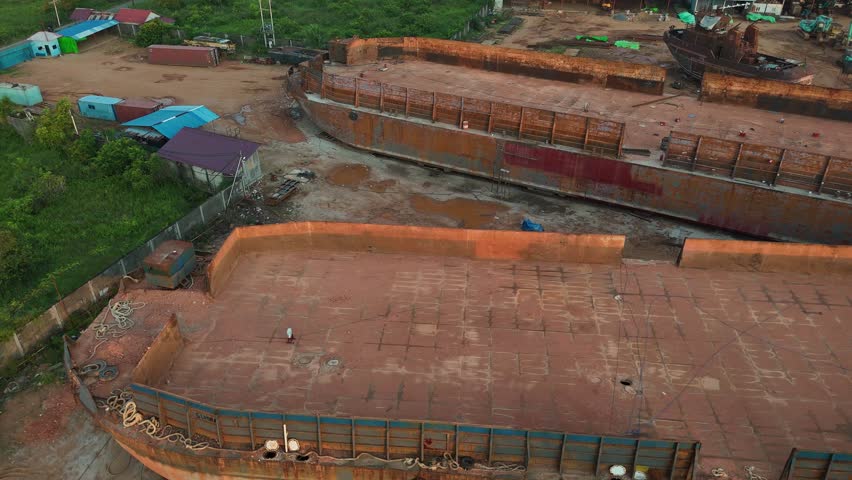 An aerial view showing a dry dock with two large, rusty steel barges side by side at the shipyard, with a crane and an old ship in the background. 