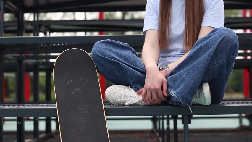 Young girl in baggy jeans sitting on metal bleachers next to skateboard. Close-up of teenage girl sitting in lotus position at stadium with skateboard. Skater resting at city sports ground