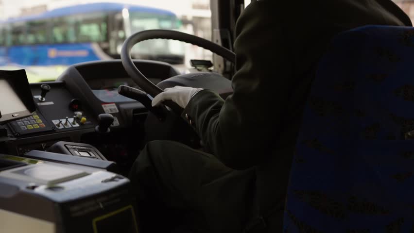 Japanese bus driver in uniform and white gloves gripping steering wheel at driver console in urban Japan.