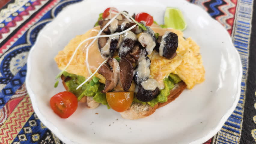 Delicious avocado egg toast served on plate in cafe. This nutritious avocado egg toast features scrambled eggs, mushrooms, cherry tomatoes, and lime on colorful tablecloth.